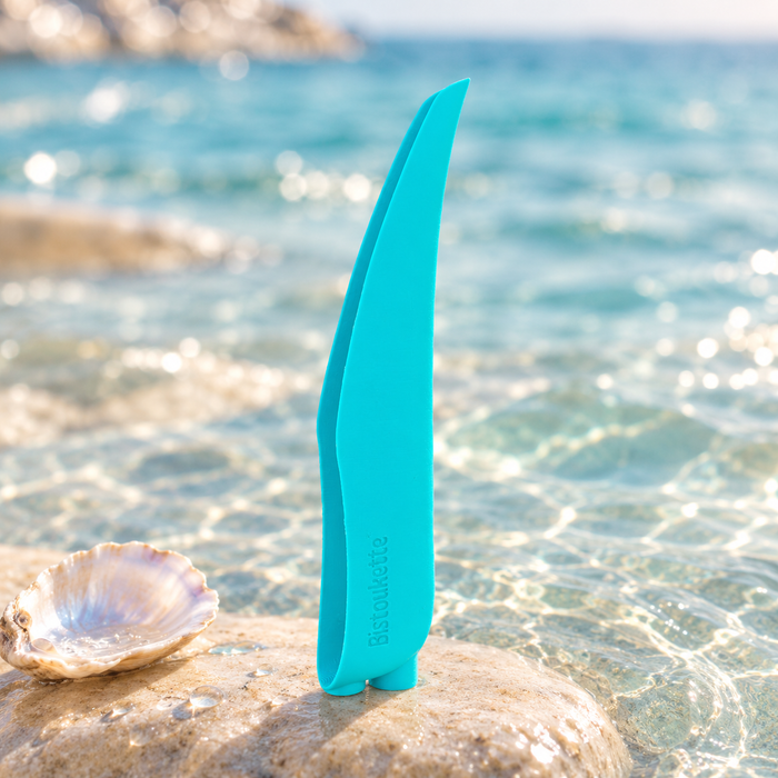 Bistoukette L'Essentielle coloris Bleue turquoise posée sur un galet dans une eau turquoise, à côté d'un coquillage nacré