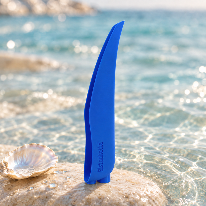 Bistoukette L'Essentielle coloris Bleue posée sur un galet au bord de la mer, à côté d'un coquillage nacré